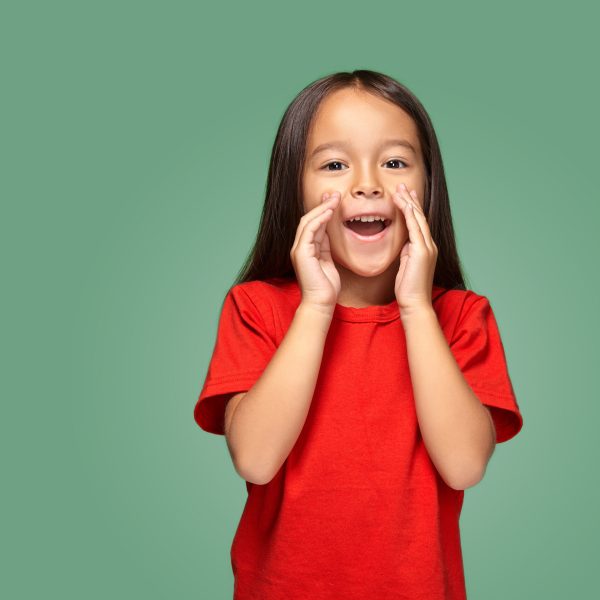 Portrait of a small pretty girl standing sideways and calling someone holding her hand near her mouth wearing a red t-shirt, green background